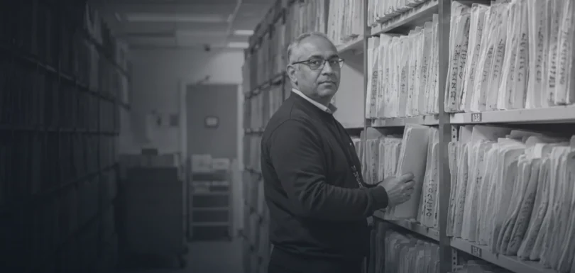 Black and white photograph of a records clerk in a hospital archive between floor-to-ceiling shelves of paper patient files
